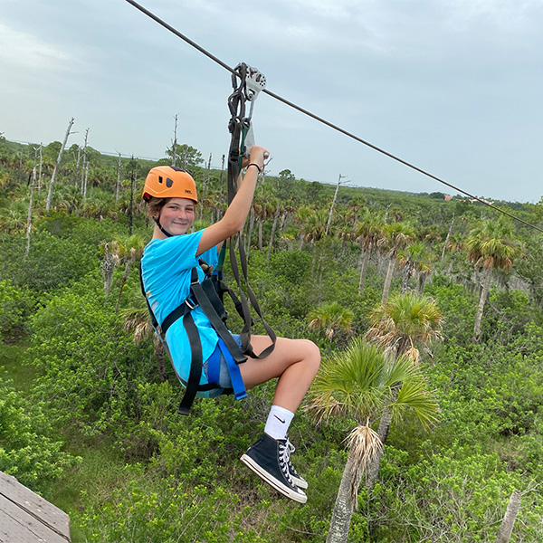 girl in blue shirt and orange helmet and black harness getting ready to zipline over the trees