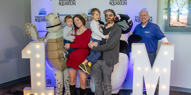 from left to right: turtle costume character, pregnant woman in a red dress holding her toddler son, man in grey jeans, black t-shirt, grey jacket, and hat holding his young son, african penguin costume character, Roger Germann CEO, standing in front of Aquarium backdrop and oversize lighted number one and oversize lighted letter M