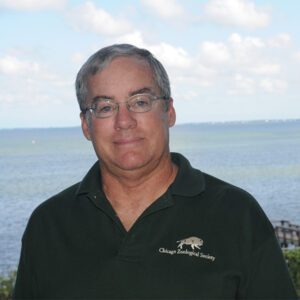 man with gray hair and glasses wearing black polo shirt in front of the ocean
