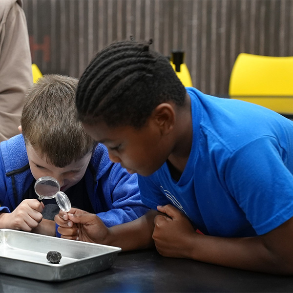 two kids wearing blue shirts using magnifying glass looking at owl droppings in a silver tray