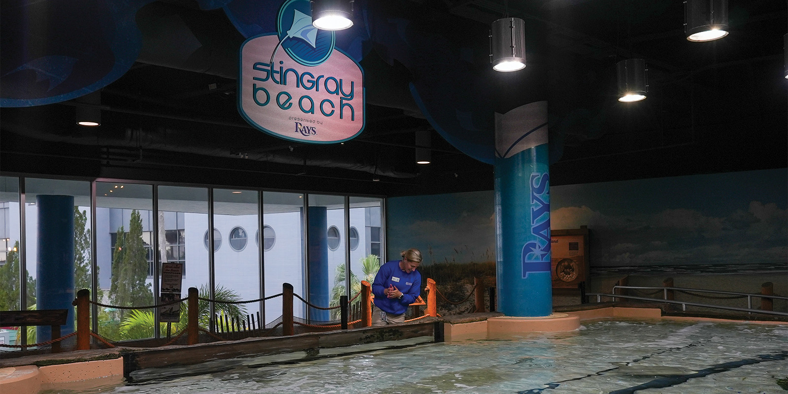 female wearing blue shirt standing under a sign which reads Stingray Beach presented by the Rays