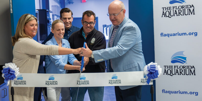 two women and three men use big blue scissors to cut a ribbon with The Florida Aquarium logo on it