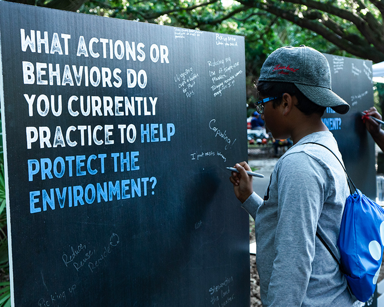 young boy writing on a sign that says 'what actions or behaviors do you currently practice to help protect the environment?'
