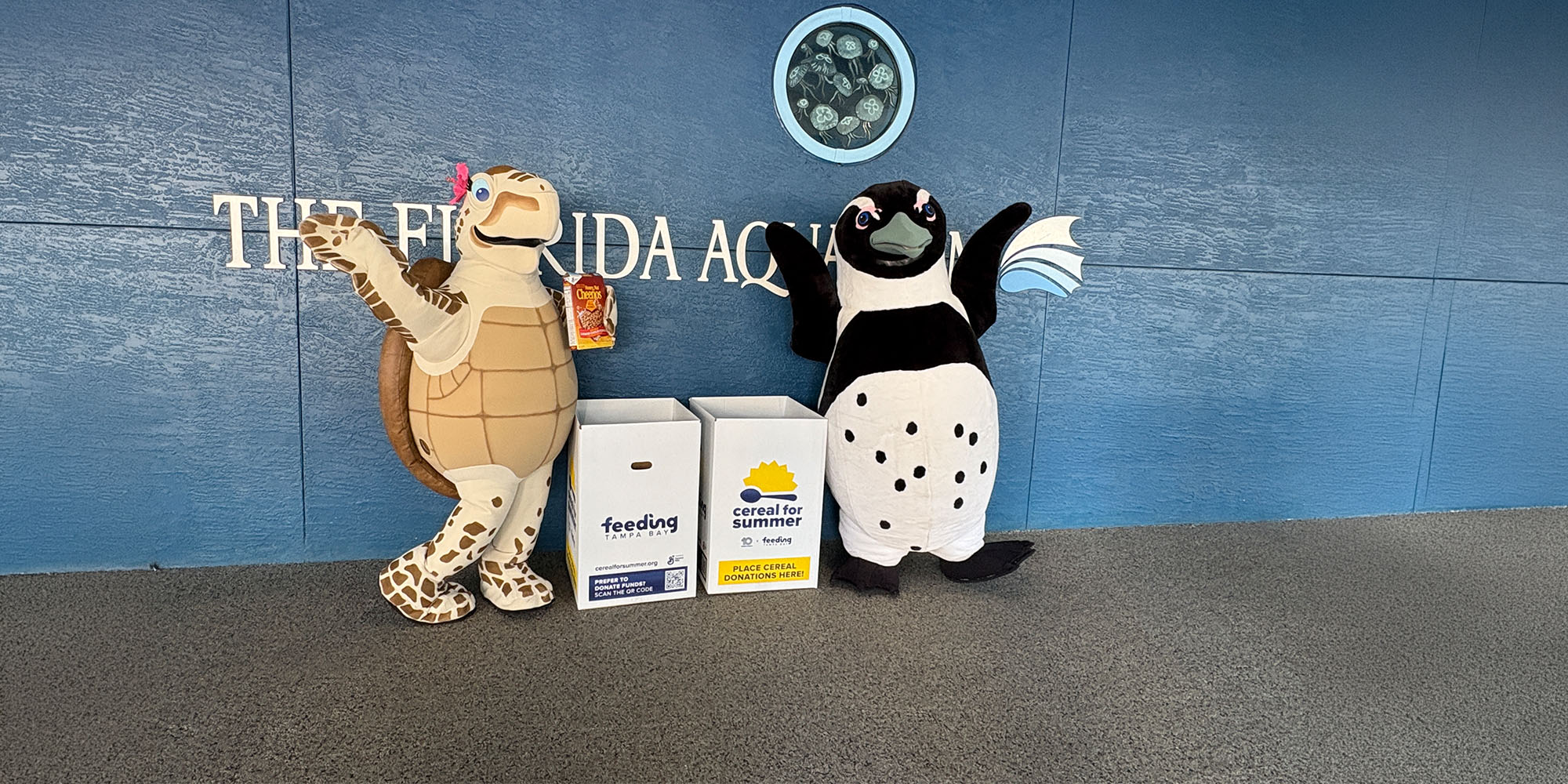 The Florida Aquarium Costume characters Tango the loggerhead turtle and Rocky the African penguin stand next to collection boxes for Feeding Tampa Bay's Cereal for Summer program outside of The Florida Aquarium in Tampa, FL.