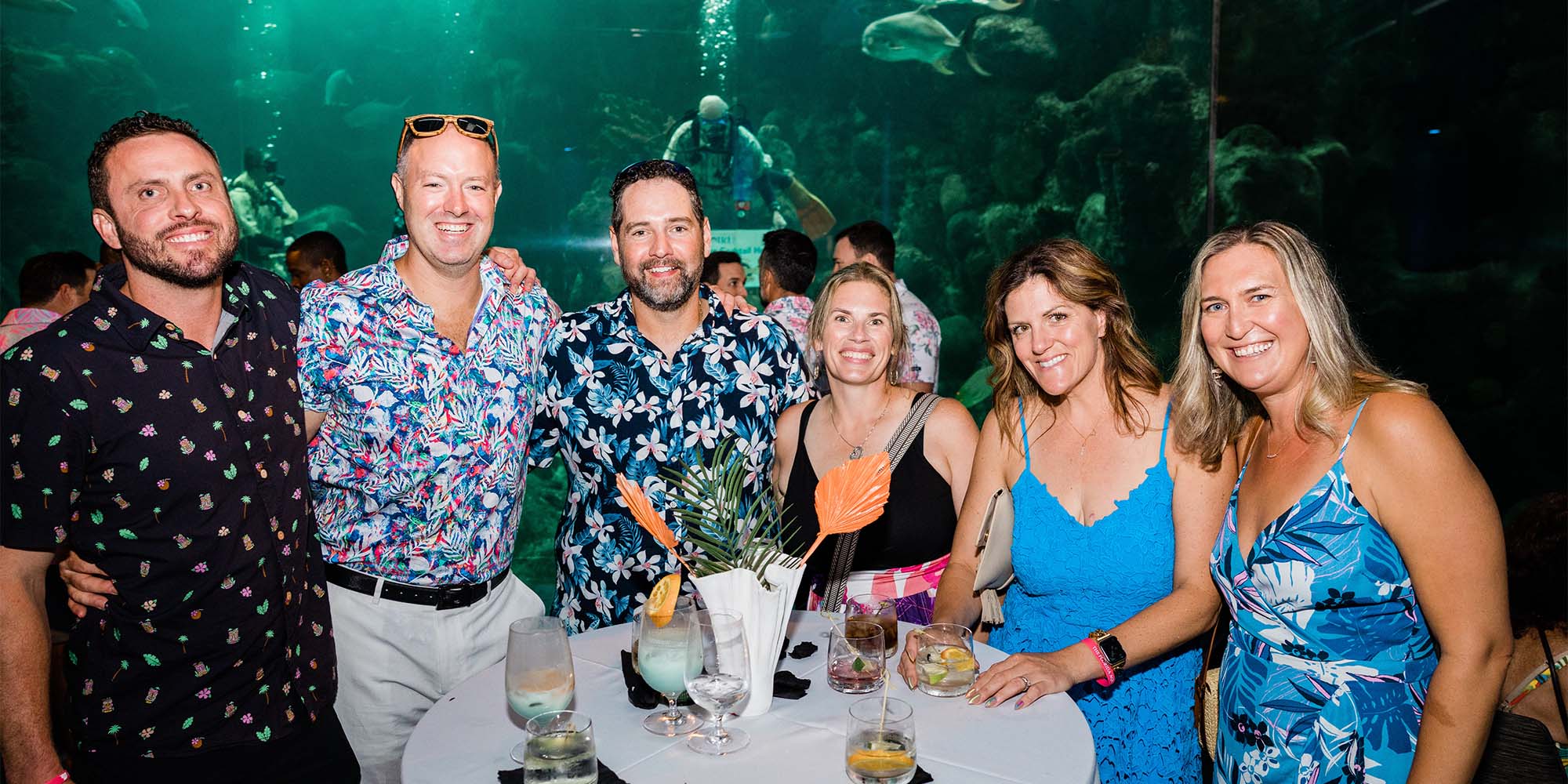 three men and three women dressed in tropical soiree clothes pose in front of The Florida Aquarium Shark Reef pan window with a cocktail table in front of them