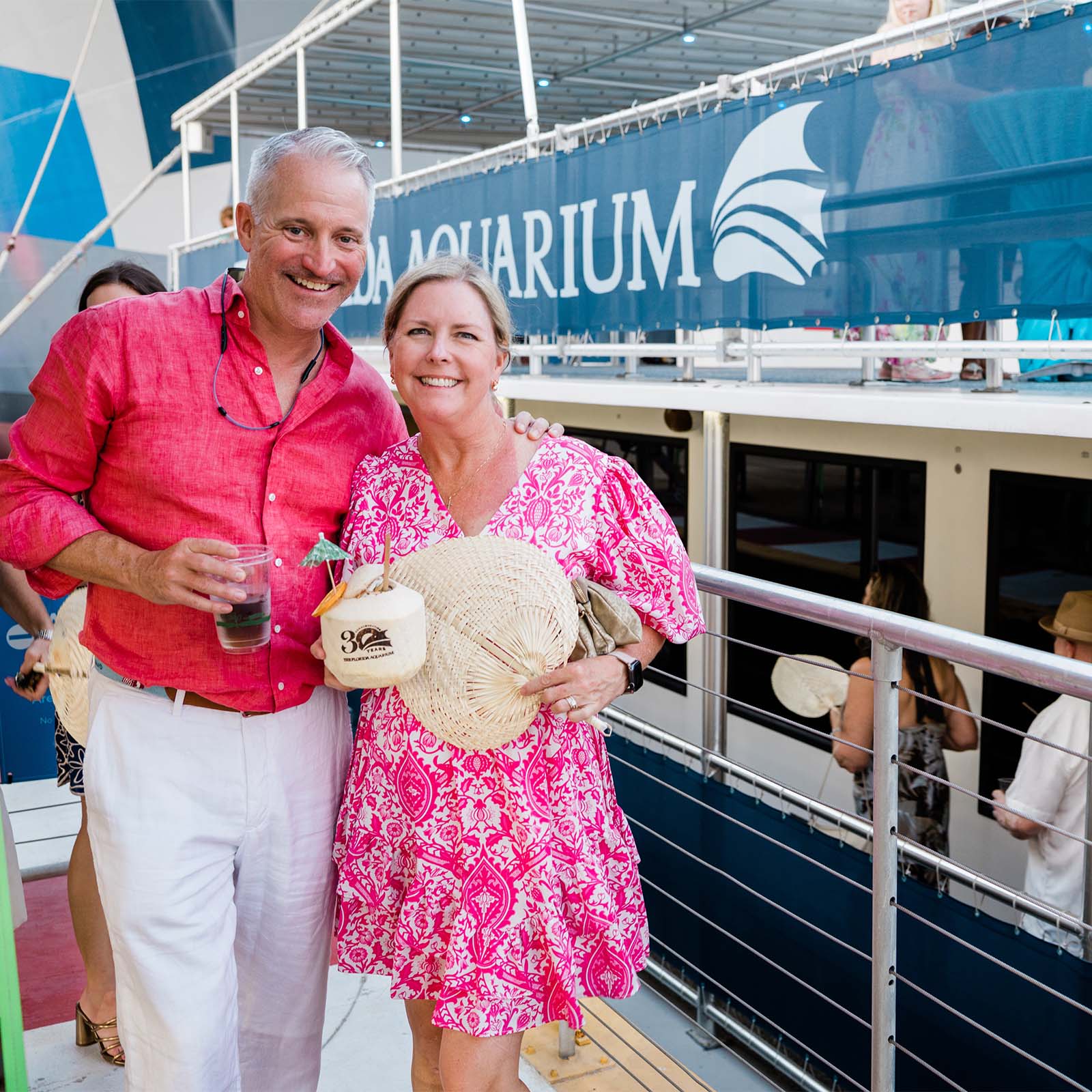 man in pink shirt and woman in pink and white printed dress stand in front of the bay spirit II boat before sunset cruise