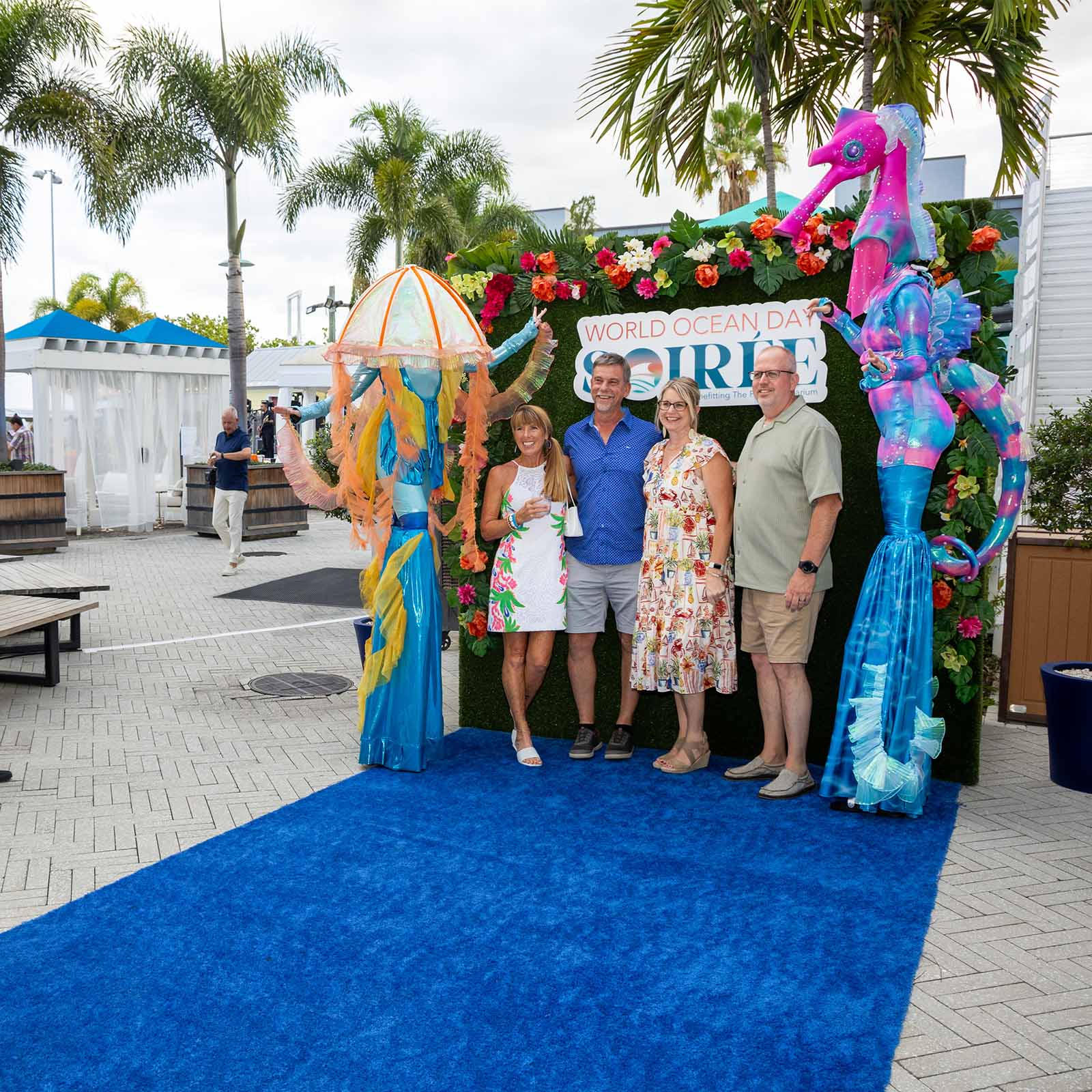 A group of women and men have their photo taken next to two stilt walkers in front of a grass wall with the World Ocean Day Soiree logo behind them