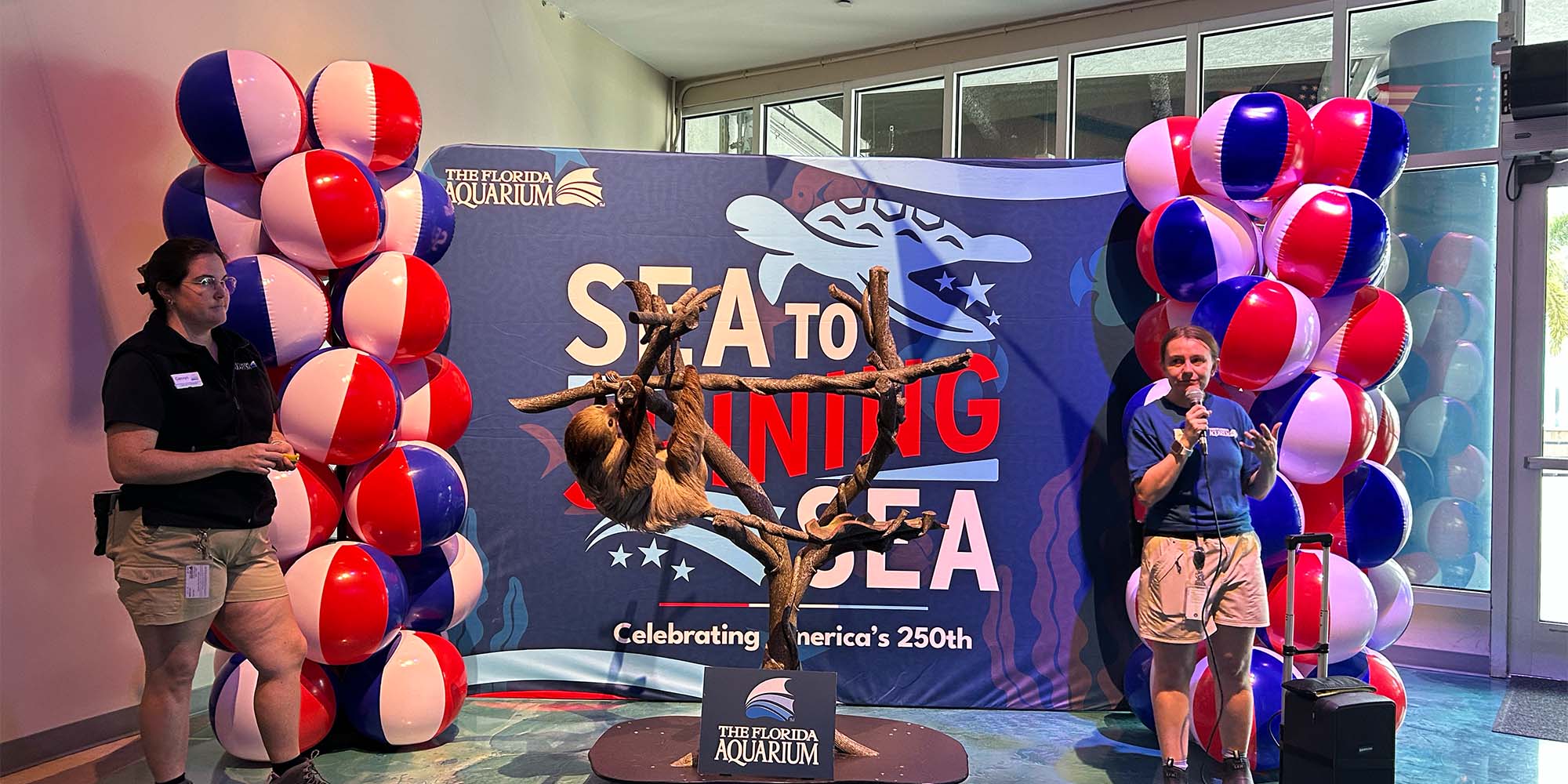 Sloth on a perch while two female employees talk to guests with Sea to Shining Sea banner in the background