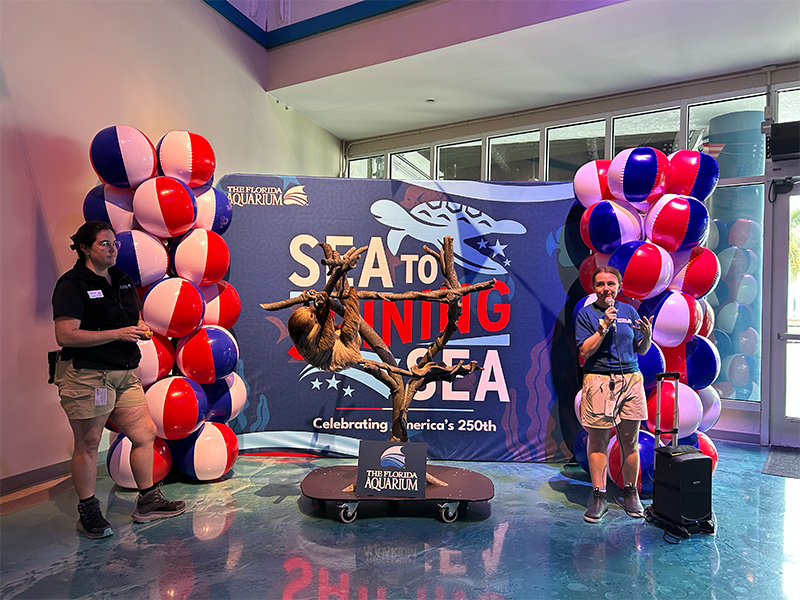 Sloth on a perch while two female employees talk to guests with Sea to Shining Sea banner in the background